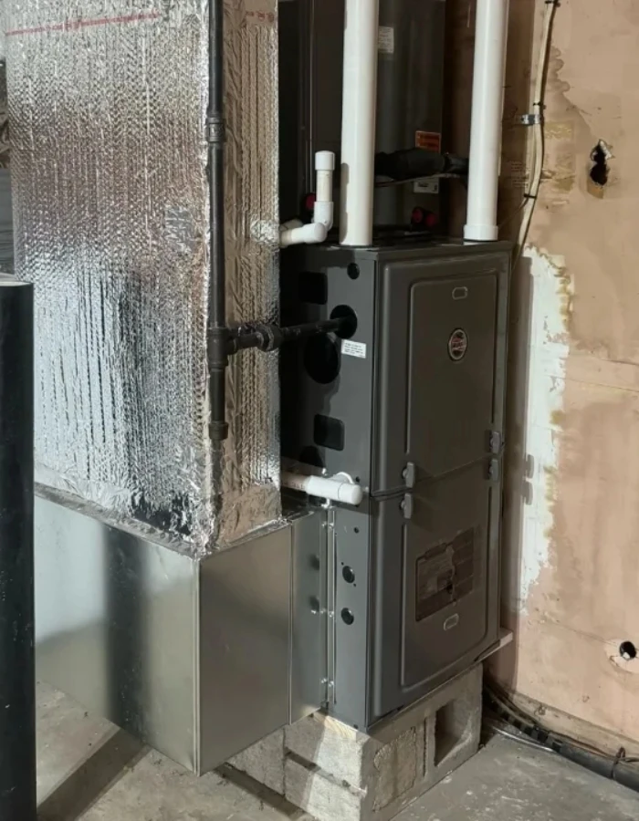 A gas furnace with combustion venting pipes (white PVC), gas line, and ductwork connections. The unit sits on concrete blocks and has a return air plenum connected at the bottom.